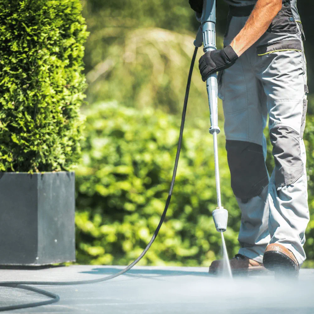 Man using a pressure washer to clean an outdoor concrete surface near a garden.